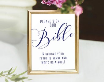 May include: A gold-framed sign with white background and navy blue text that reads "Please sign our Bible. Highlight your favorite verse and write us a note!"