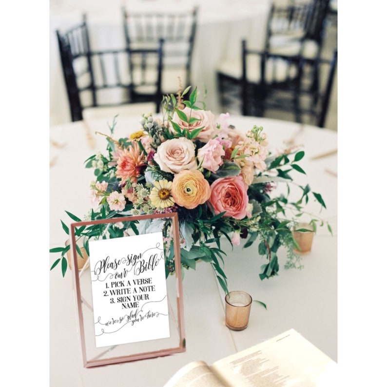 May include: A white sign in a gold frame with the text "Please Sign Our Bible" and instructions to pick a verse, write a note, and sign your name. The sign is on a table with a bouquet of pink, peach, and yellow flowers.