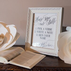 May include: A white framed sign with the text "Please sign our Bible" and instructions to pick a verse, write a note, and sign your name. A closed Bible is on a wooden table.