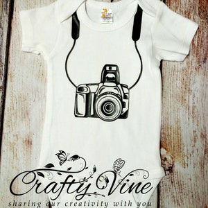 May include: White baby onesie with a black and white camera graphic and black straps. The onesie has the text "Crafty Vine" and "sharing our creativity with you" in black script.