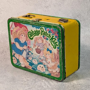 Puede incluir: Fiambrera vintage de Cabbage Patch Kids. La fiambrera es amarilla con un borde verde y presenta ilustraciones de Cabbage Patch Kids. Tiene un cierre y un asa de metal. La fiambrera rectangular tiene un aspecto ligeramente desgastado.