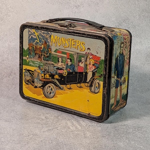 May include: Vintage metal lunchbox featuring a colorful illustration of the Munster family in their car, with the text "The Munsters". The lunchbox has a black handle and a black border.