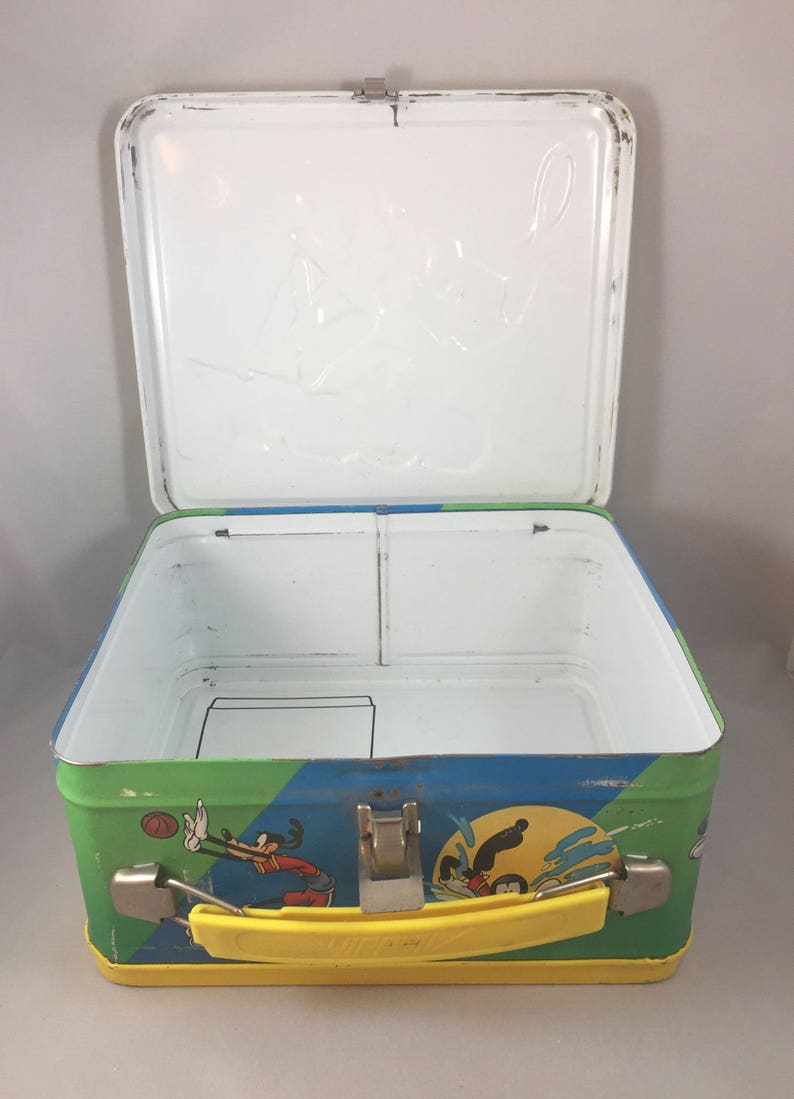 May include: A vintage metal lunchbox featuring a colorful design of Mickey Mouse and Goofy playing sports. The lunchbox has a yellow handle and a white interior with a divider.