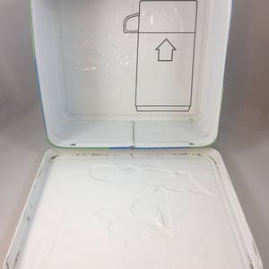 May include: A yellow, blue, and green metal lunchbox with a white interior. The lunchbox has a latch and a handle. The inside of the lunchbox has a molded design of a cartoon character.