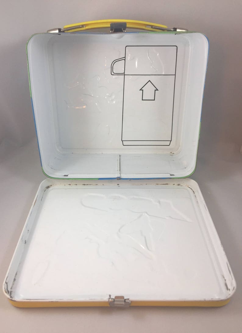 May include: A yellow, blue, and green metal lunchbox with a white interior. The lunchbox has a latch and a handle. The inside of the lunchbox has a molded design of a cartoon character.