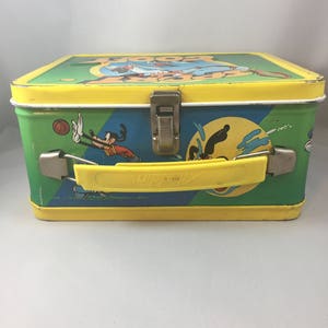 May include: A vintage metal lunchbox featuring a cartoon image of Goofy playing various sports. The lunchbox is yellow, green, and blue with a yellow handle. The text "Goofy" is printed on the top of the lunchbox.