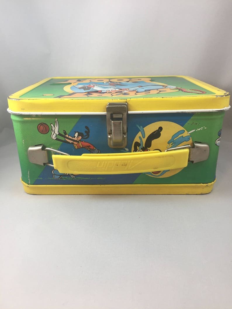 May include: A vintage metal lunchbox featuring a cartoon image of Goofy playing various sports. The lunchbox is yellow, green, and blue with a yellow handle. The text "Goofy" is printed on the top of the lunchbox.