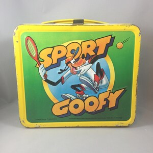 May include: A yellow metal lunchbox with a green background featuring Goofy playing tennis. The lunchbox has the words "Sport Goofy" on it.