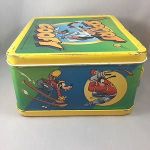 May include: A yellow metal lunchbox with a green, blue, and yellow design. The lunchbox features a cartoon image of Goofy and Mickey Mouse skiing and playing baseball. The text "Sport Goofy" is printed on the top of the lunchbox.