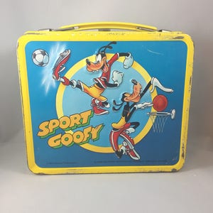May include: Yellow metal lunchbox featuring Goofy and a cartoon version of himself playing sports. The lunchbox has the words "Sport Goofy" on the front.