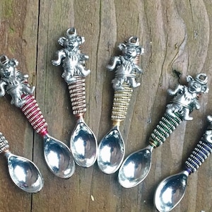 May include: Six silver spoons with colorful wire wrapped handles. Each spoon features a small bear figurine on the top. The bears are all in different poses.
