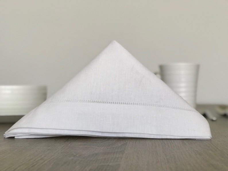 May include: A white linen napkin folded into a triangle shape, with a subtle stitched border. The napkin is sitting on a wooden table.