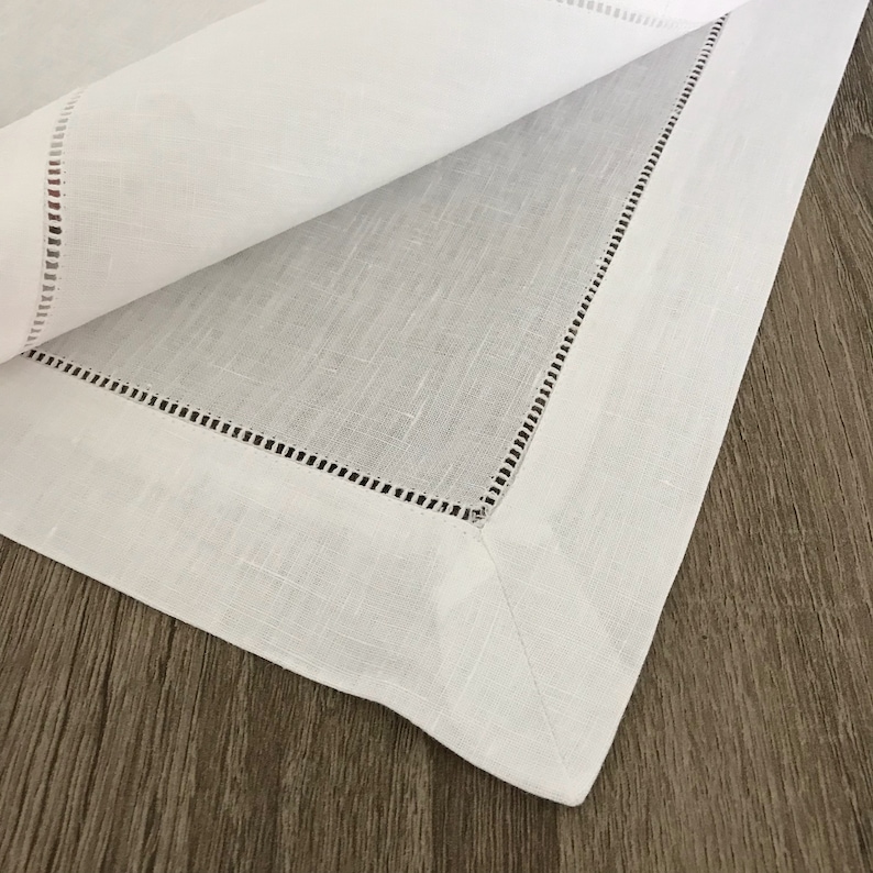 May include: White linen placemat with a delicate, stitched border. The placemat is folded in half, revealing the texture of the linen.