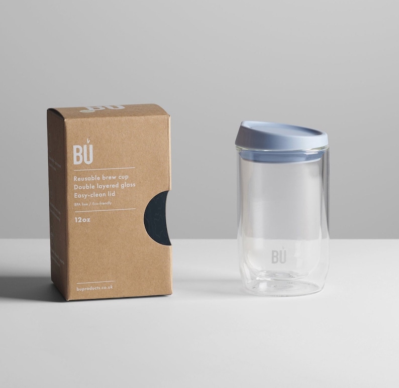 May include: A reusable glass brew cup with a double-layered design and a light blue lid. The cup is 12oz and has the text "BU" on the side. The cup is sitting on a white surface next to a brown box with the text "BU" and "Reusable brew cup" on it.
