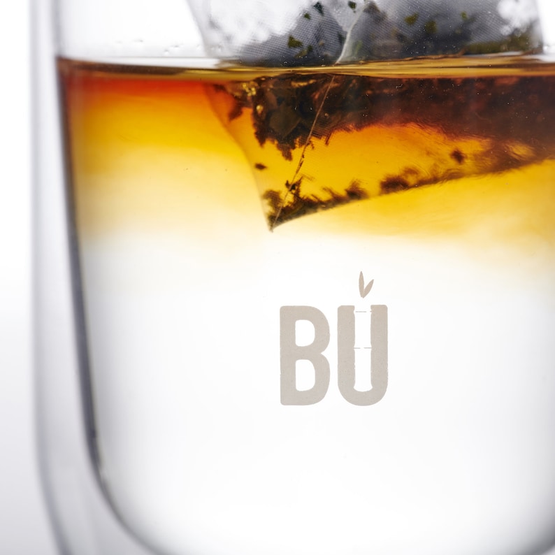 May include: A clear glass cup with a tea bag submerged in the water. The tea bag is filled with loose leaf tea and is partially submerged in the water. The water is a light amber colour. The word "B&Uuml;" is printed on the bottom of the glass.