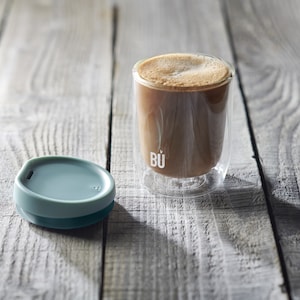May include: A clear glass coffee mug with a double wall design and a teal silicone lid. The mug has the word "B&Uuml;" printed on the side. The mug is filled with coffee and is sitting on a wooden surface.