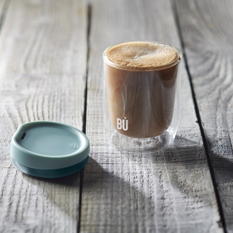 May include: A clear glass coffee mug with a double wall design and a teal silicone lid. The mug has the word "B&Uuml;" printed on the side. The mug is filled with coffee and is sitting on a wooden surface.