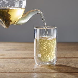 May include: A clear glass teapot containing green tea leaves is pouring into a double-walled glass cup with the word "BU" printed on the side. The cup is filled with light yellow tea.