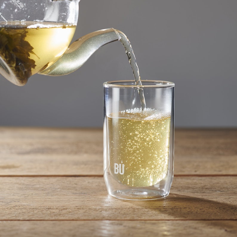 May include: A clear glass teapot containing green tea leaves is pouring into a double-walled glass cup with the word "BU" printed on the side. The cup is filled with light yellow tea.