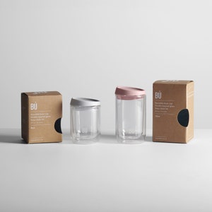 May include: Two reusable glass coffee cups with lids. The cup on the left has a white lid and the cup on the right has a pink lid. Both cups are labelled "B&Uuml;" and are sitting on a white surface. The text on the box reads "Reusable brew cup, Double layered glass, Easy-clean lid, 12oz."