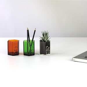 Könnte beinhalten: Drei Glasbehälter, zwei grün und einer braun, mit den Zahlen "A45", "G30" und "G30" eingeprägt. Die grünen Behälter enthalten Bleistifte und der braune Behälter enthält eine kleine Sukkulente.