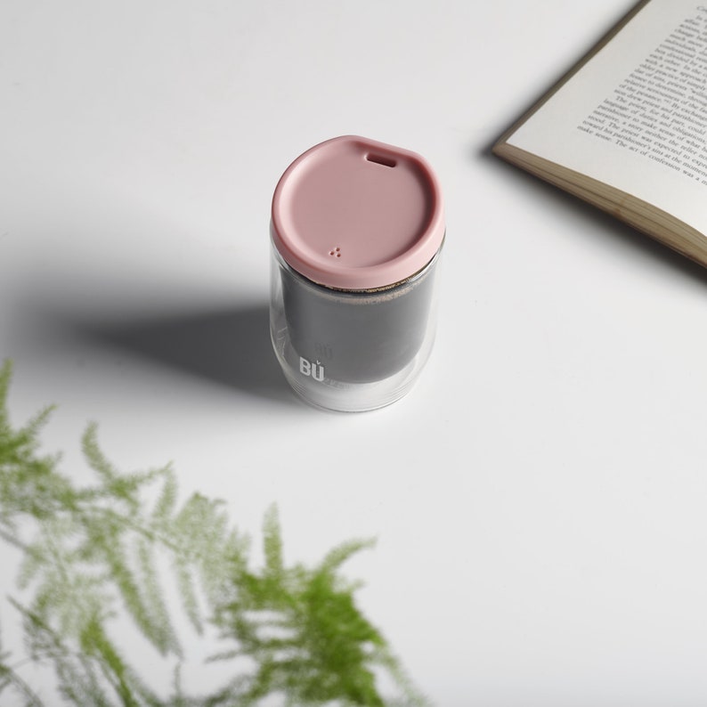 May include: A double-walled glass mug with a pink lid. The mug has a black silicone sleeve and the text "BU" printed on the side.