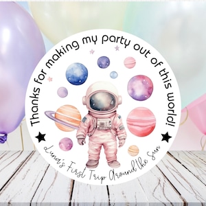 May include: Round white sticker with a cartoon astronaut in a pink spacesuit. The sticker has a black star and the text "Thanks for making my party out of this world! Luna's First Trip Around the Sun".