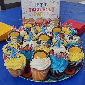 May include: A platter of cupcakes decorated with dragon and taco themed toppers. The cupcakes are frosted in yellow, white, and blue. A sign in the background reads "Let's Taco 'Bout Party".