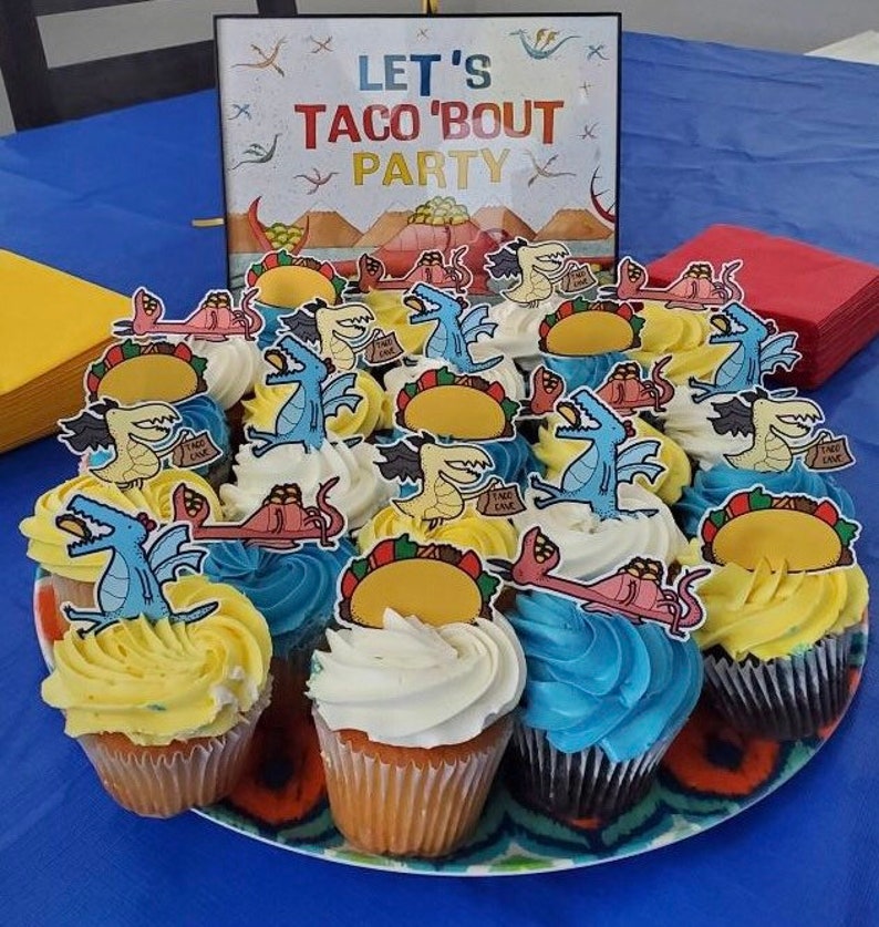 May include: A platter of cupcakes decorated with dragon and taco themed toppers. The cupcakes are frosted in yellow, white, and blue. A sign in the background reads "Let's Taco 'Bout Party".