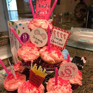 K&ouml;nnte beinhalten: Ein mehrst&ouml;ckiger Cupcake-St&auml;nder mit rosa glasierten Cupcakes, jeder mit rosa Streuseln und einer rosa Kerze. Dekorative Aufs&auml;tze enthalten Texte wie "I'm a mouse, duh!" und "You go, Glen Coco!"
