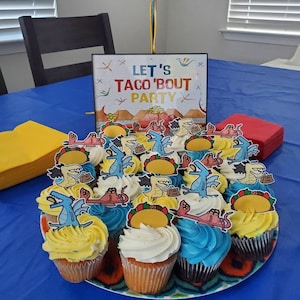 May include: A blue table with a sign that says "Let's Taco 'Bout Party" and a plate of cupcakes decorated with dinosaurs and tacos. The cupcakes are frosted in blue, yellow, and white.