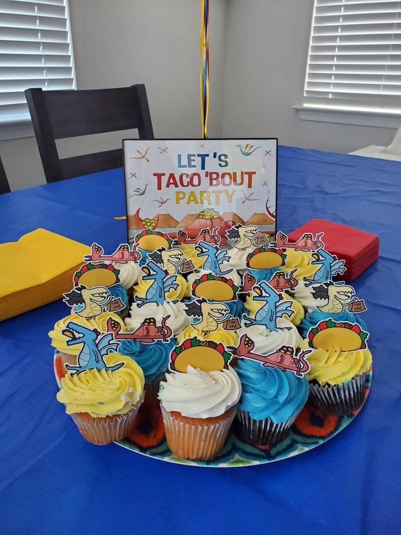 May include: A blue table with a sign that says "Let's Taco 'Bout Party" and a plate of cupcakes decorated with dinosaurs and tacos. The cupcakes are frosted in blue, yellow, and white.