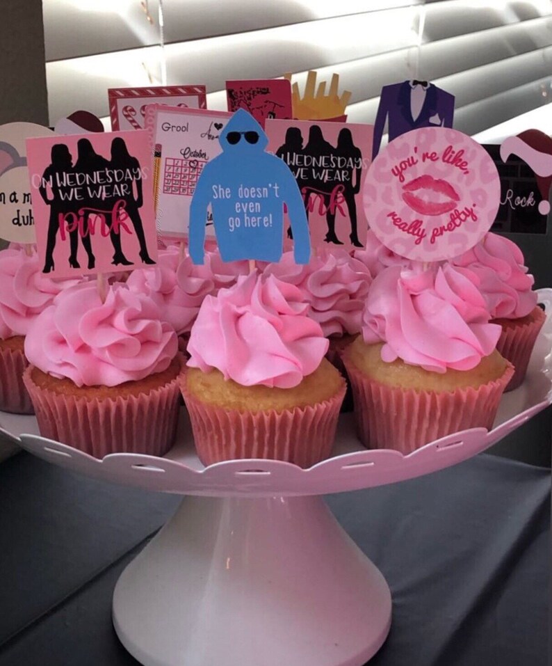 K&ouml;nnte beinhalten: Eine Sammlung von Cupcakes mit rosa Zuckerguss, pr&auml;sentiert auf einem wei&szlig;en, mehrst&ouml;ckigen Servierst&auml;nder. Die Cupcakes sind mit verschiedenen Toppern verziert, darunter Silhouetten, eine blaue Kapuzenpullover-Grafik mit Text und ein rosa Lippen-Design mit Text.