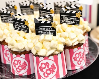 May include: A tray of cupcakes decorated with popcorn and clapperboard toppers. Each cupcake has a white marshmallow topping and is in a pink and white popcorn-themed wrapper. The clapperboards read "Emerson's 6th Birthday."