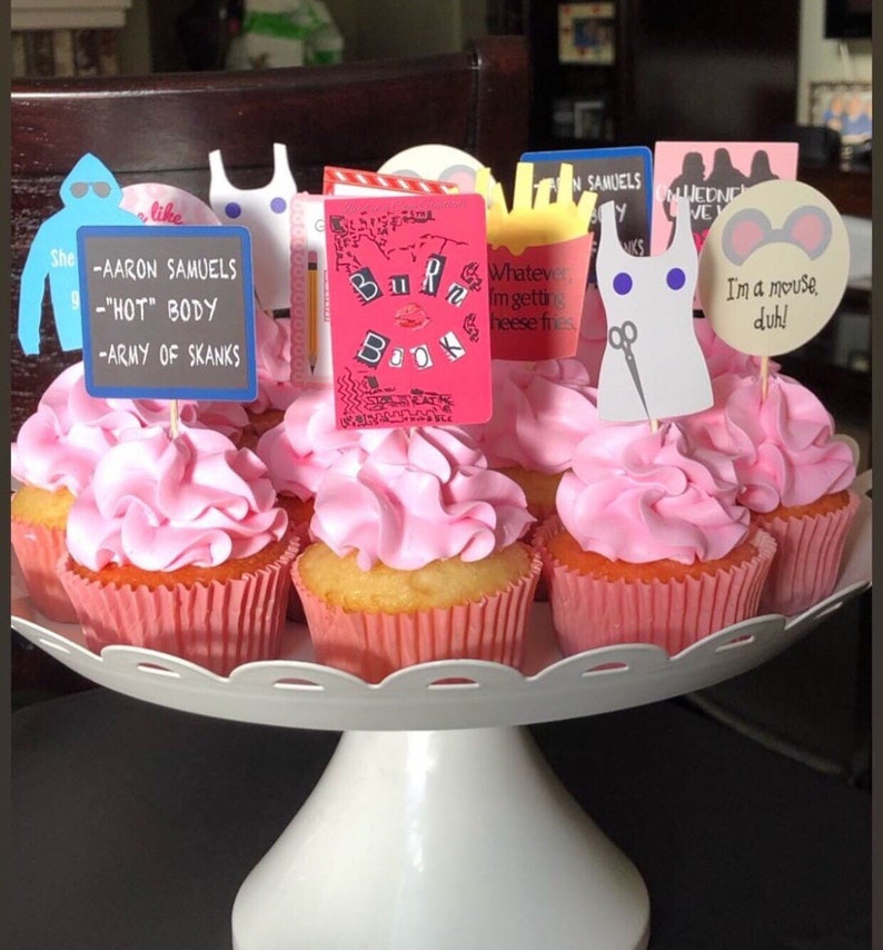 K&ouml;nnte beinhalten: Ein wei&szlig;er, mehrst&ouml;ckiger Kuchenst&auml;nder mit Cupcakes mit rosa Zuckerguss und Dekorationen. Die Dekorationen zeigen Text und Bilder, darunter ein "Burn Book" und den Satz "I'm a mouse, duh!". Die Cupcakes sind in rosa Papierf&ouml;rmchen.