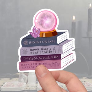 May include: A sticker featuring a stack of books with titles like "Hexes for Exes" and "Moon Magic & Manifestations." A crystal ball and amethyst crystals sit atop the books. The sticker has a white border and is held by a hand.