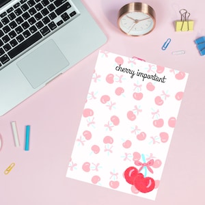 May include: A white notepad with a pink cherry pattern and the text "cherry important" printed on it. The notepad is on a pink surface with a laptop, a clock, and office supplies.