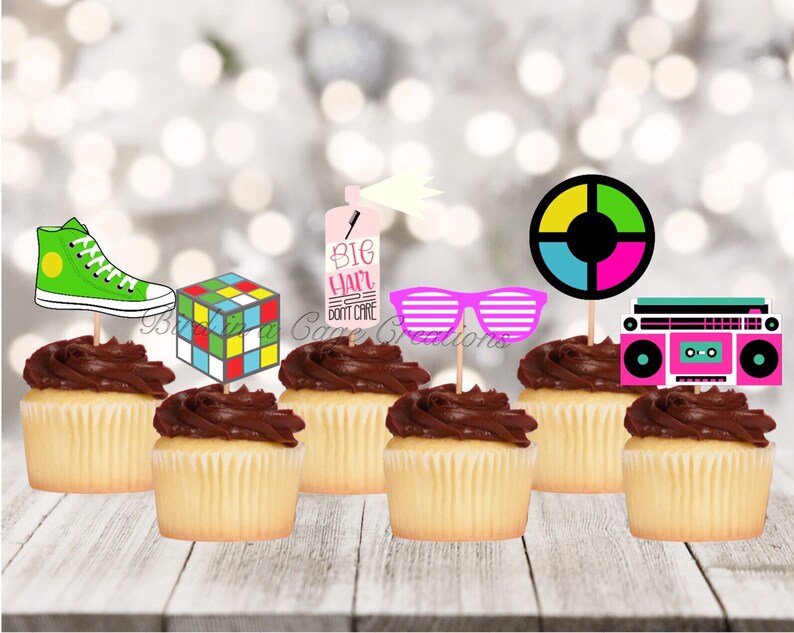 May include: A set of 8 cupcake toppers featuring 80s themed designs. The toppers include a green sneaker, a Rubik's cube, a pink and blue boombox, a pair of pink sunglasses, a spray can with the text "Big Hair Don't Care", and a colourful circle with a black centre.