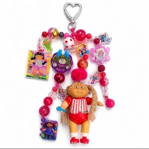 May include: A decorative keychain featuring a heart-shaped clasp, pink and red beads, and multiple Cabbage Patch Kids charms. The centerpiece is a small doll with blonde braided hair wearing a red and white striped shirt and red shorts.