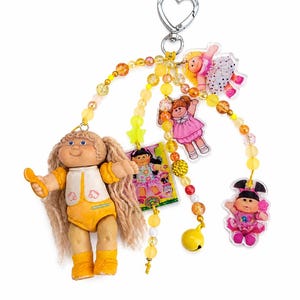 May include: A Cabbage Patch Kids keychain featuring a small doll with blonde hair and yellow outfit. The keychain includes a heart-shaped clasp, beaded strands, and several small, colorful Cabbage Patch Kids charms. The overall color scheme is yellow and orange.