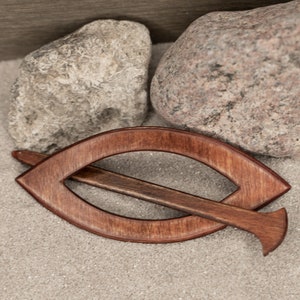 May include: A wooden hair stick with a curved, oval-shaped design. The stick is brown and has a smooth finish. It is resting on a bed of sand.
