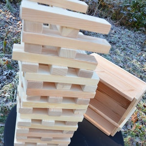 May include: A tall tower constructed from rectangular light-colored wooden blocks stacked in alternating horizontal layers, accompanied by a matching open-top wooden storage crate positioned to the right side of the stacked game tower.