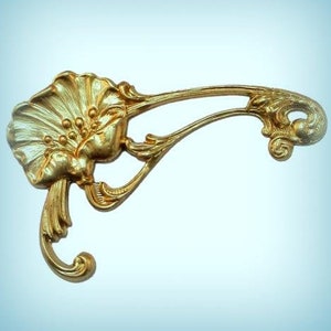 May include: Ornate gold-colored decorative hardware with a floral design. The piece features a stylized flower with detailed petals and a curved, flowing arm. The hardware is likely for furniture or decorative purposes.