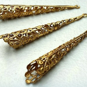 K&ouml;nnte beinhalten: Drei verzierte, goldfarbene Metall-Kegel-Schmuckteile. Jedes St&uuml;ck weist aufwendige Filigranarbeiten und eine kleine Schlaufe am spitz zulaufenden Ende auf. Die Schmuckteile werden auf hellem Hintergrund pr&auml;sentiert.