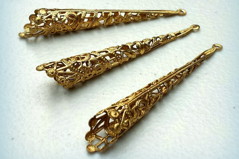 K&ouml;nnte beinhalten: Drei verzierte, goldfarbene Metall-Kegel-Schmuckteile. Jedes St&uuml;ck weist aufwendige Filigranarbeiten und eine kleine Schlaufe am spitz zulaufenden Ende auf. Die Schmuckteile werden auf hellem Hintergrund pr&auml;sentiert.