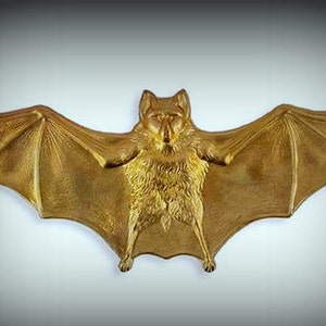 May include: A gold-colored metal bat figurine with outstretched wings. The bat has a detailed, textured body and a realistic appearance.