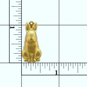 May include: A gold-colored metal charm in the shape of a rabbit. The rabbit is sitting upright with its ears perked up. The charm is about 1 inch tall.