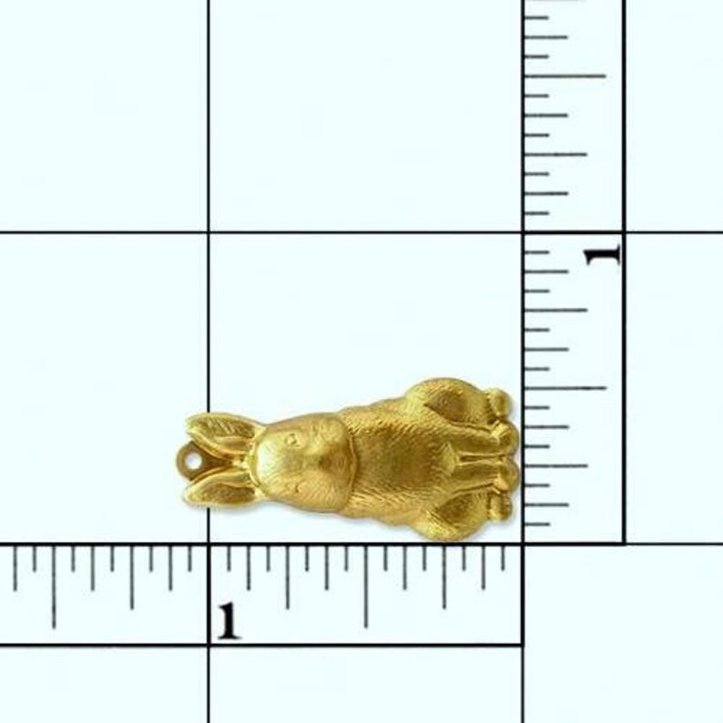 May include: A gold-colored metal rabbit charm, approximately 1 inch long, lying on a white surface with a ruler for scale.