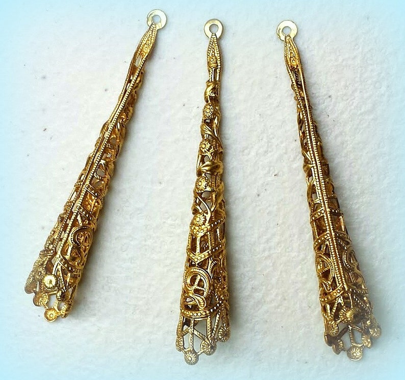 K&ouml;nnte beinhalten: Drei goldfarbene, konisch geformte Schmuckteile aus Filigran. Jeder Konus hat ein offenes Design mit komplizierten floralen und geometrischen Mustern. Die Teile haben oben eine kleine Schlaufe zur Befestigung an anderen Schmuckkomponenten. Die Konen sind etwa 5 cm lang.