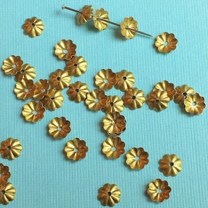 May include: A collection of gold-colored metal flower-shaped embellishments, scattered on a light blue surface. The embellishments have a scalloped edge and a small hole in the center.